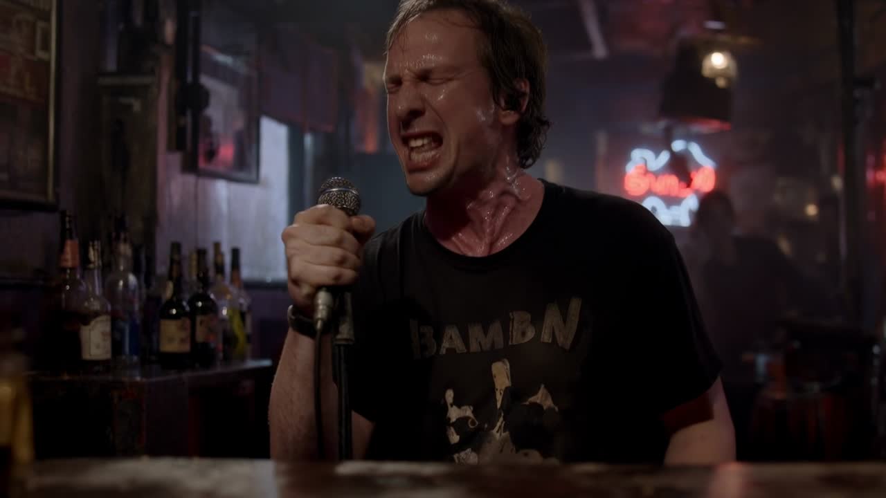 Inspirando, cantante maschile in maglietta scura che stringe il microfono, proiettando ed esibendosi in un bar con insegna al neon