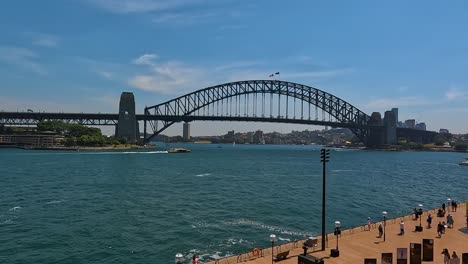 Sydney-Harbour-Bridge-overlooking-bustling-promenade,-sunny-day,-blue-water