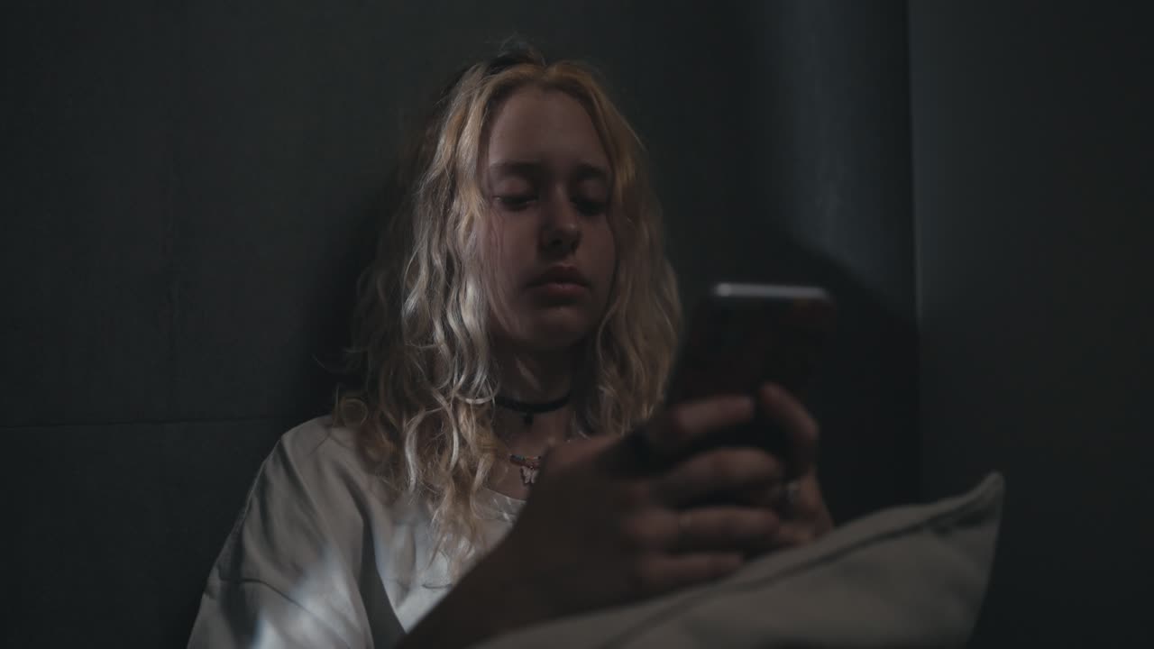 una chica rubia con una camiseta blanca se apoya contra una pared gris y mira las redes sociales antes de irse a la cama tarde en la noche