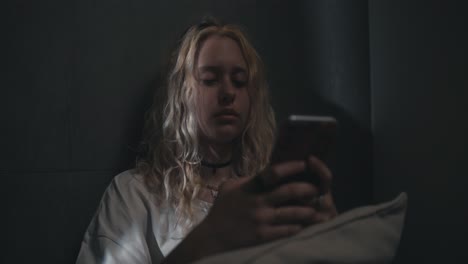 A-blonde-girl-in-a-white-T-shirt-leans-against-a-gray-wall-and-watches-social-networks-before-going-to-bed-late-at-night