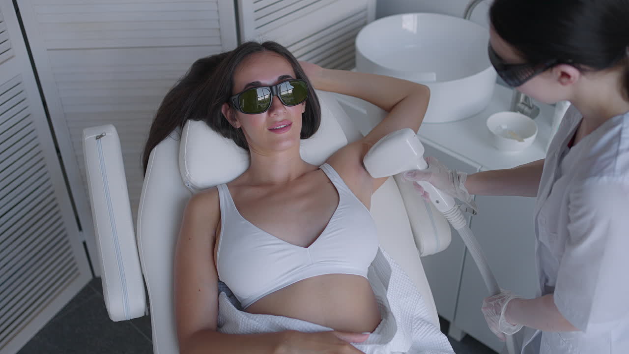 미용사 는 의료 소파 에 누워서 미용실 에서 웃는 매력적 인 섹시 한 여자 의 겨드랑이 레이저 제거 를 하고 있다. 미용사 는 환자 의 겨드랑이 레이저 제거 를 한다. 탈모 절차
