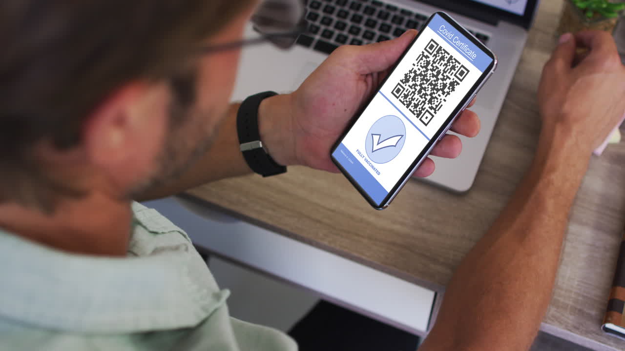 Man at desk holding smartphone with covid vaccination certificate, tick and qr code on screen