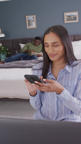 video vertical de una mujer biracial en el dormitorio usando un teléfono inteligente y sonriendo, pareja en el fondo