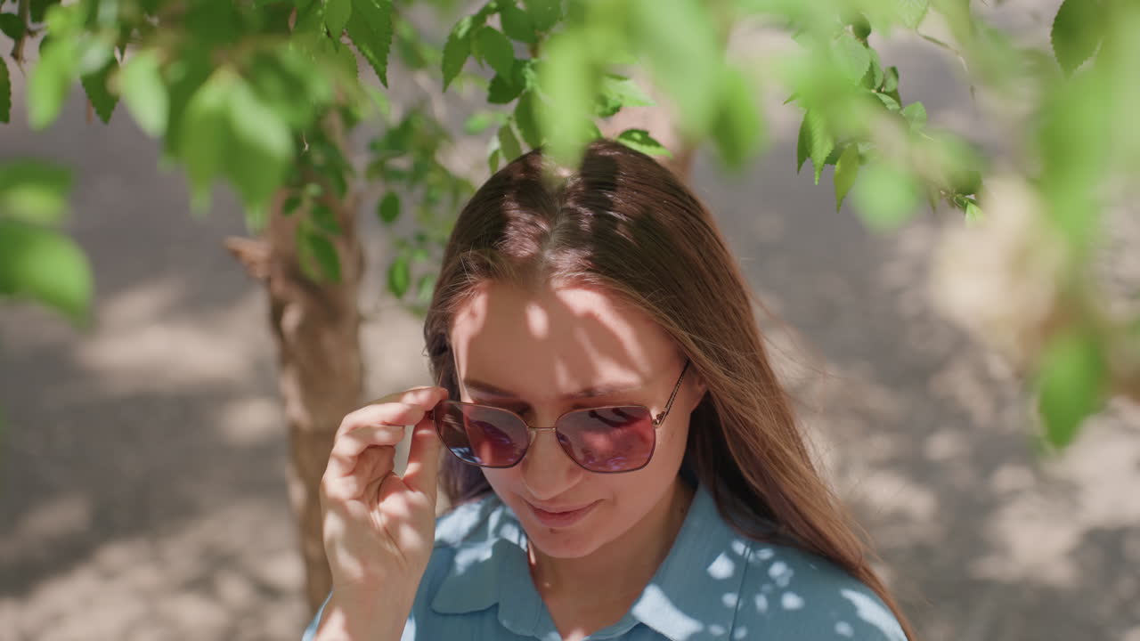 Энергичная женщина улыбается под зеленой листвой: оживленная сцена, на которой женщина греется в лучах яркого солнца; радостная женщина с удовольствием демонстрирует свои солнцезащитные очки на фоне яркой лиственной природы.