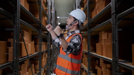vista lateral de ingeniero masculino asiático con casco de seguridad de pie y gritando meta celebrando trabajando en el almacén con estantes llenos de mercancías de entrega