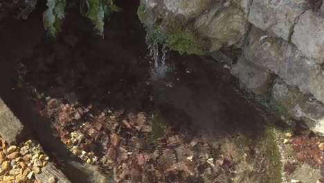 Wasser,-Das-Aus-Einer-Alten-Steinmauer-In-Einen-Kleinen-Teich-Fließt