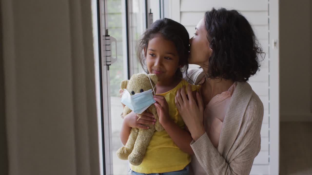madre y hija hispanas abrazándose en la ventana sosteniendo un oso de peluche con máscara facial