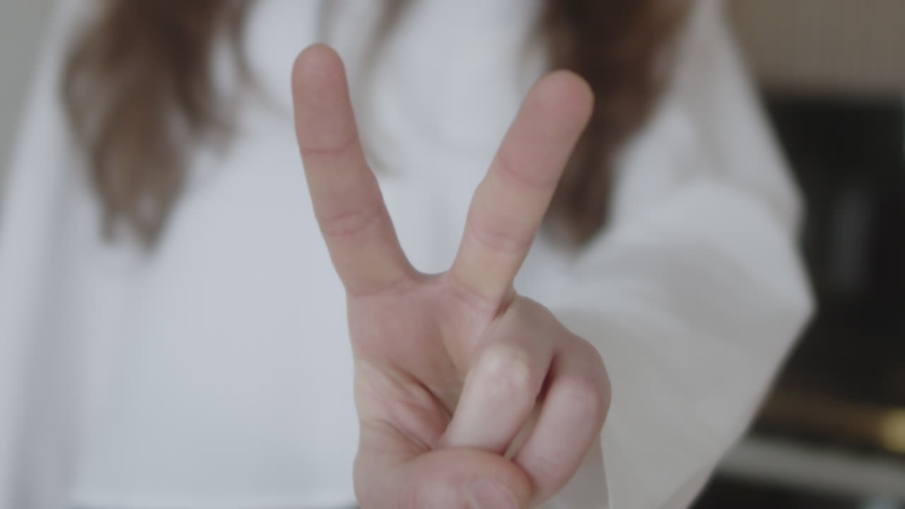 beautiful slow motion close-up shot of a young Caucasian woman's fingers raising her index and middle finger forming a “v” and “love and peace” in a white shirt at home in natural light.