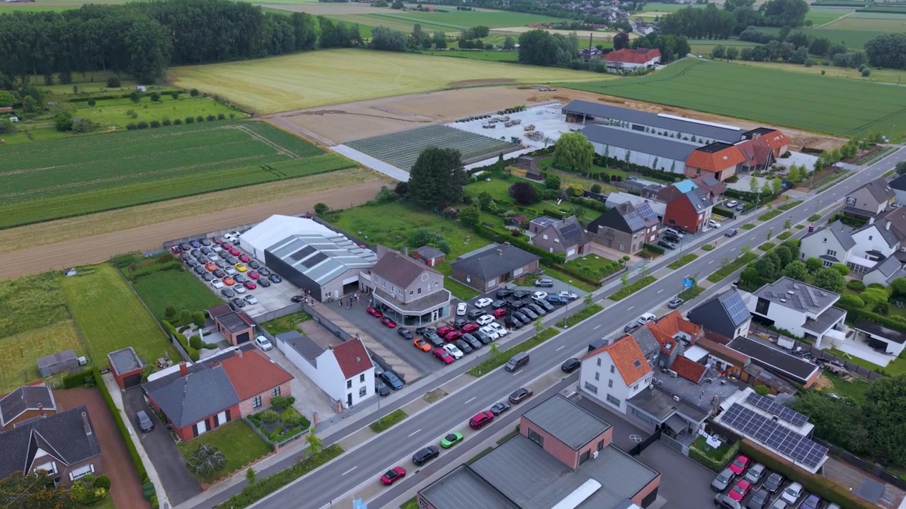 Luchtfoto van een autobedrijf omringd door landbouwgrond en woongebouwen, toont geparkeerde voertuigen, moderne faciliteiten, toegangswegen en een landelijk landschap overdag