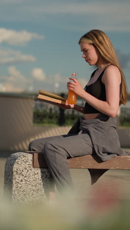 mujer sentada en un banco bebiendo de una botella de jugo mientras continúa leyendo un libro, la escena presenta un puente superior y un fondo borroso con postes de lámpara