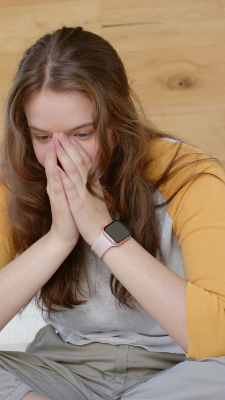 Video verticale: Donna seduta a casa con le mani che si coprono, che guarda in basso, con orologio rosa e maglietta color senape