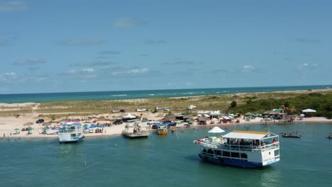 Rotating-aerial-drone-shot-of-a-large-tourist-transport-ship-arriving-at-the-dock-of-the-beautiful-Restinga-beach-where-the-large-Curimataú-river-meets-the-sea-near-Barra-do-Cunhaú,-Brazil