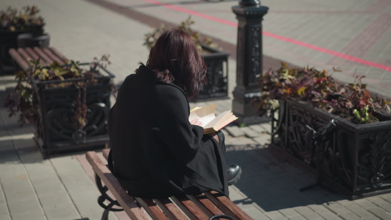 야외에 앉아서 책을 읽고 얼굴을 는 머리카락으로 새로운 페이지를 넘기고, 장식 비에 있는 꽃이 바람에 부드럽게 흔들리는 평화로운 도시 환경.