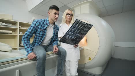 woman doctor radiologist explains the results of mri scanning for young male patient, showing the snapshot with images, observing and analyzing mri scan in modern clinic beside modern closed-type mri