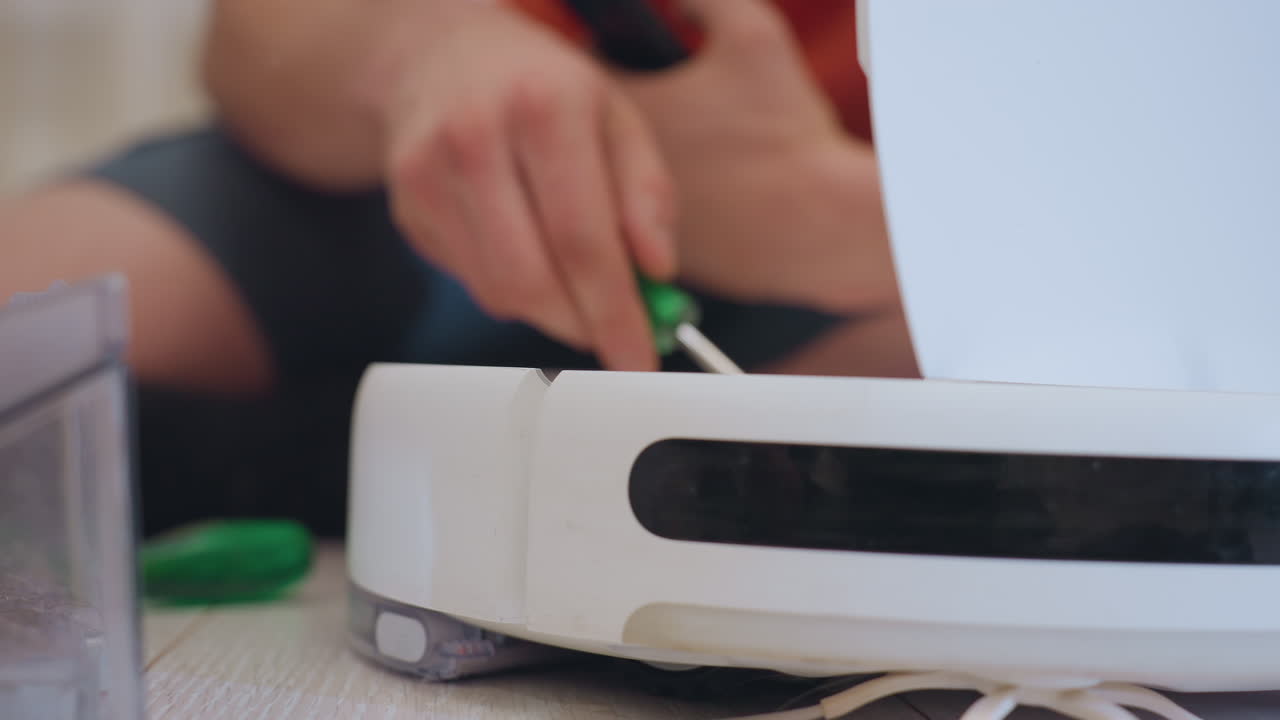 Close up of vacuum technician in orange shirt seated on floor holding tool while inspecting robot vacuum cleaner with open lid showing detailed repair process