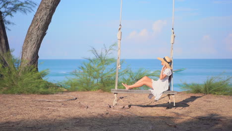 a young woman in a blue sundress and floppy straw sun hat gently sways back and forth on a rustic rope and wood swing