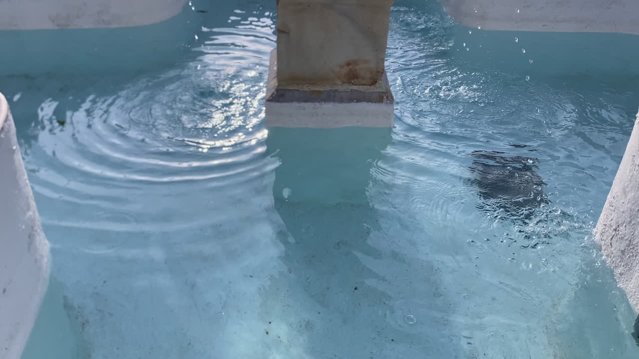 Vanddråber falder uafbrudt ned i en forvitret stenfontæne, hvilket skaber ringe på den klare blå overflade og reflekterer lys.