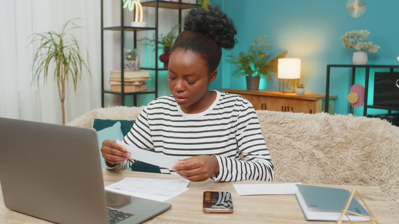Donna afroamericana che gestisce finanze e budget sul tavolo con calcolatrice, bollette e laptop, lavorando da casa
