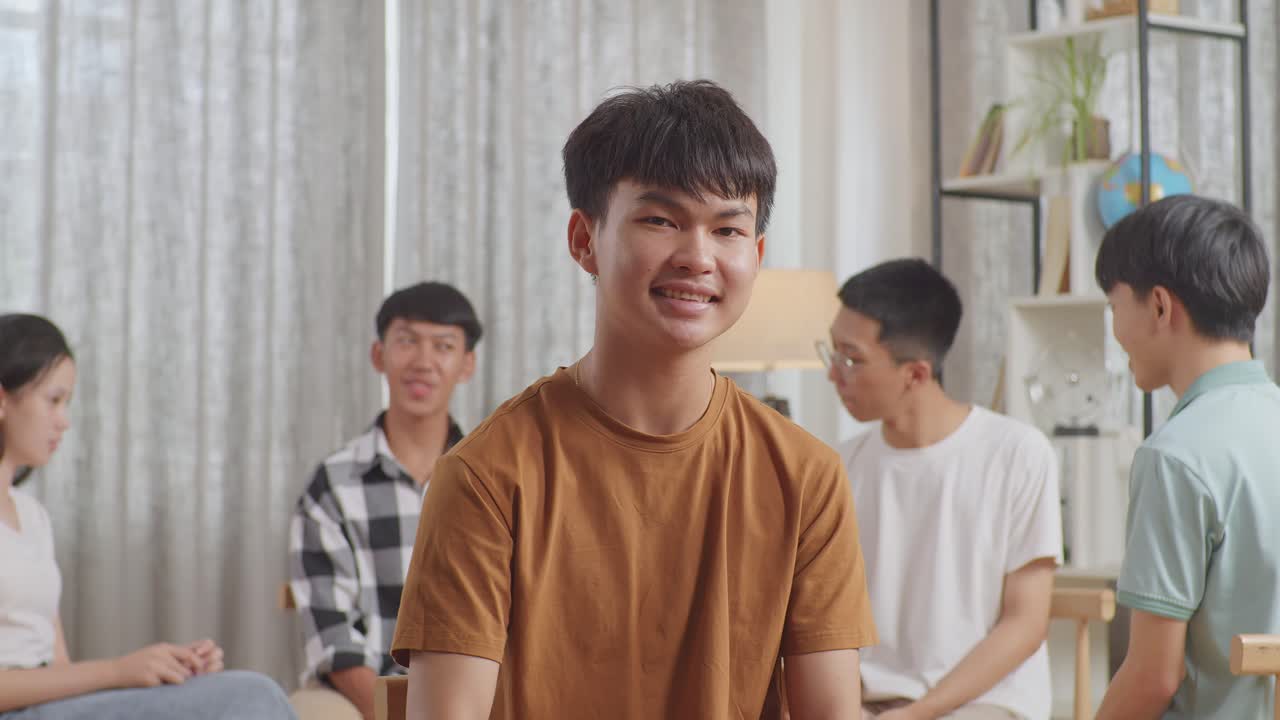 카메라에 미소 짓는 소년의 클로즈업. 아시아 십대 그룹이 의자에 앉아서 집에서 논의하는 원을 형성합니다.
