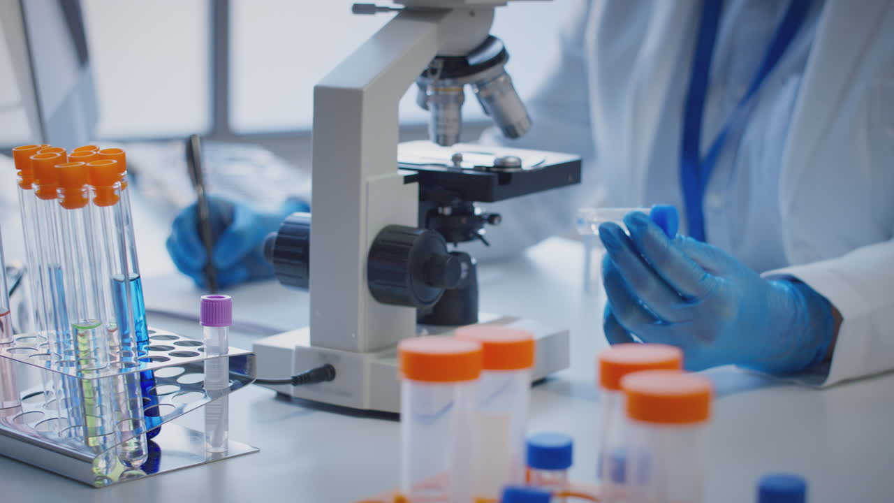 Close Up Of Lab Worker Conducting Research Using Microscope Holding Test Tube Labelled Alpha