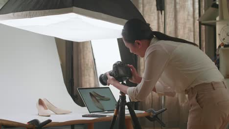 asian female photographer comparing photos on a laptop to camera while taking photos of women's shoes in home studio