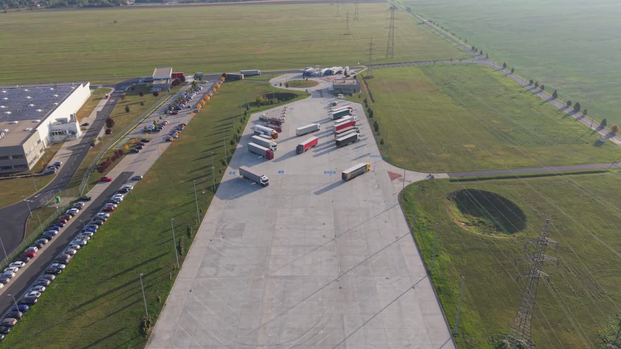 A drone view of a large truck parking area adjacent to a logistics warehouse. Several semi-trailers and trucks are parked on the concrete lot surrounded by green fields and industrial roads