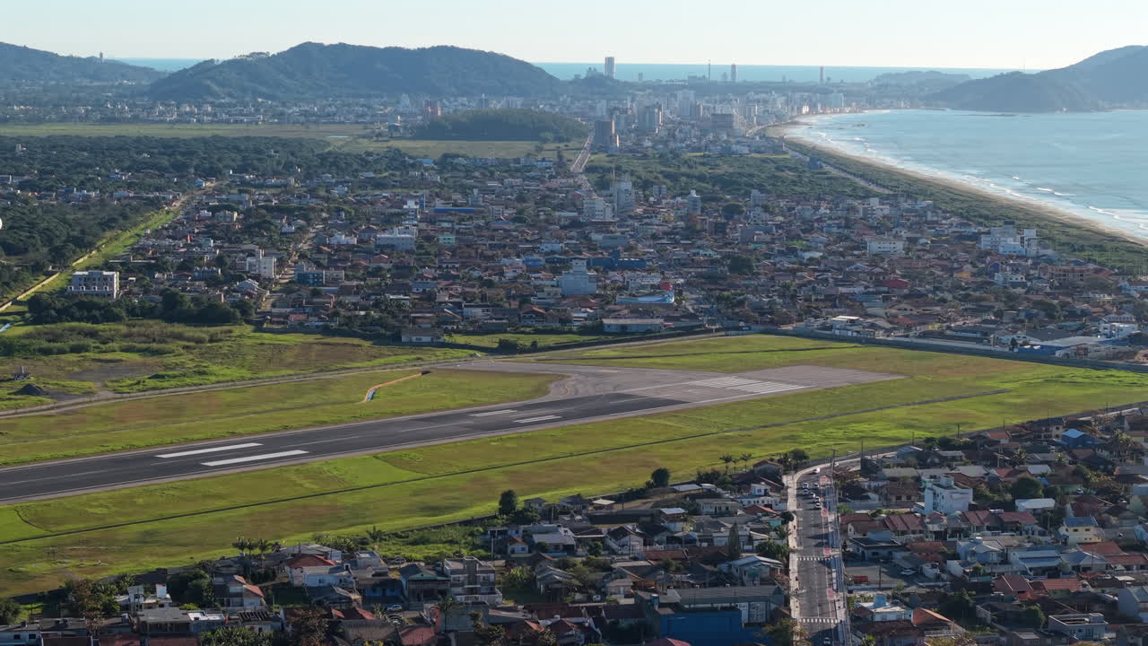 Drone view of Navegantes Airport showing the full runway surrounded by fields, urban areas, coastline, sea, and mountains in the background