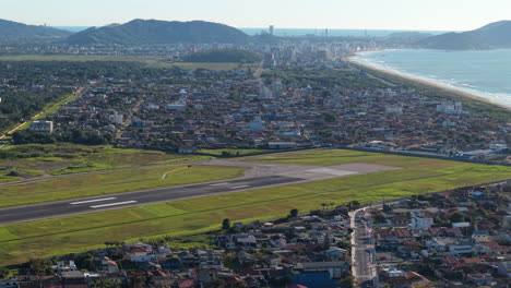 drone view of navegantes airport showing the full runway surrounded by fields, urban areas, coastline, sea, and mountains in the background