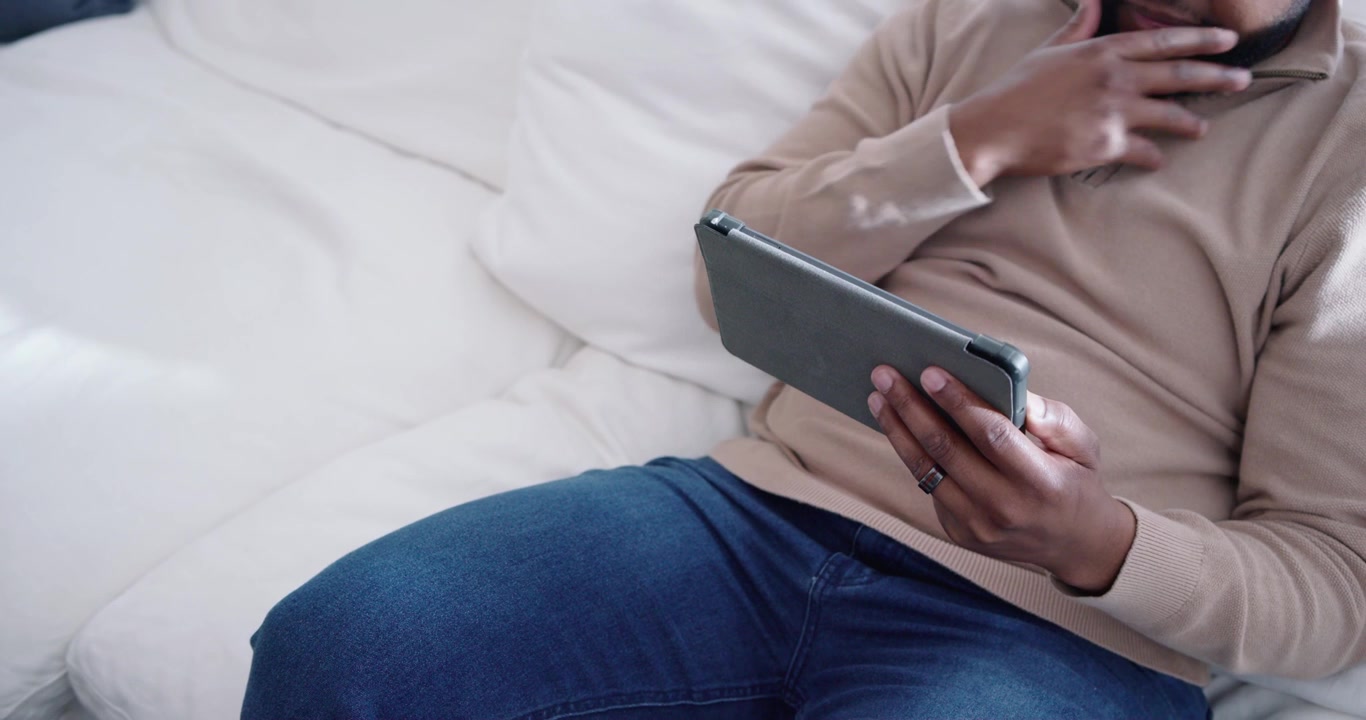 Man holding tablet, tapping and swiping screen, adjusting sweater collar and browsing on white sofa