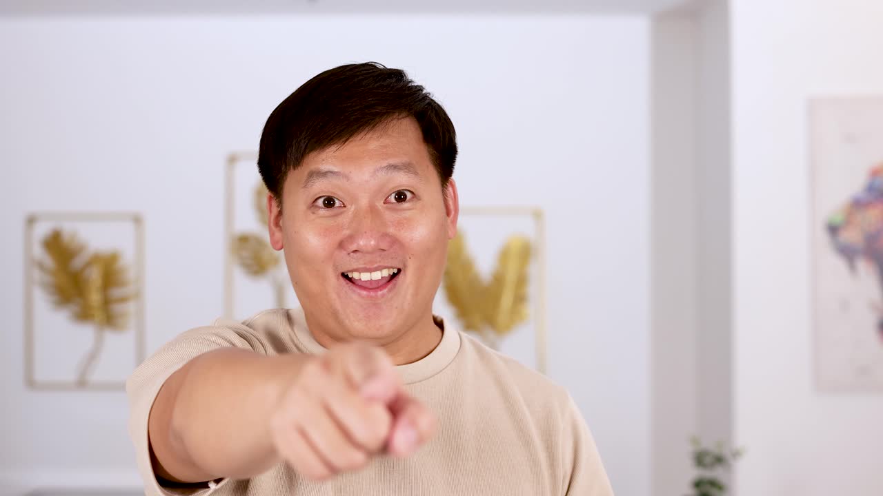 Asian man in casual beige shirt points and smiles at camera in a well-lit modern living room, with steady camera and cheerful mood