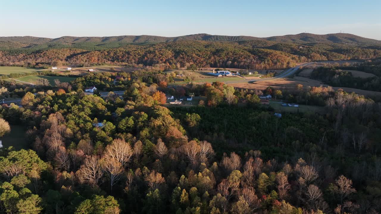 Aerial rising wide shot of peaceful American rural landscape with rolling hills, autumn forests, open fields and scattered farmhouses under clear sky during golden hour. Virginia, USA in fall