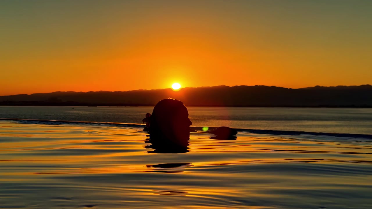 Persona che si rilassa in una piscina a sfioro al tramonto con vista sulle montagne e acqua calma