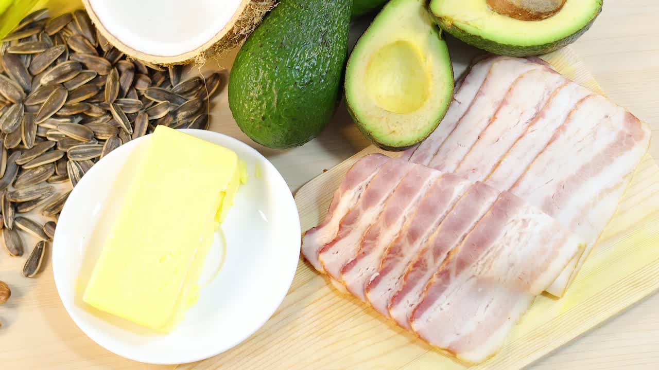 A selection of high-fat foods including butter, bacon, avocados, and seeds arranged on a wooden surface