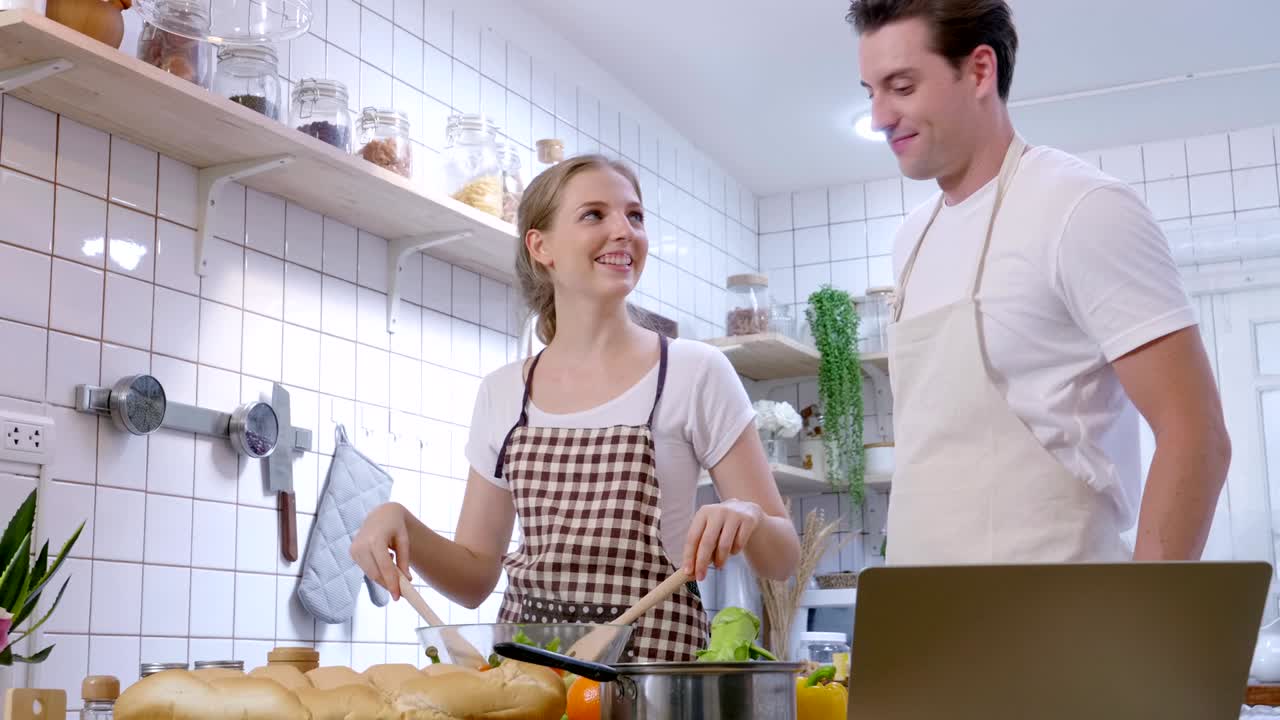 pareja caucásica cocinando comida en la cocina mientras ve un tutorial de video en una computadora portátil, aprendiendo tutorial de cocina en las redes sociales