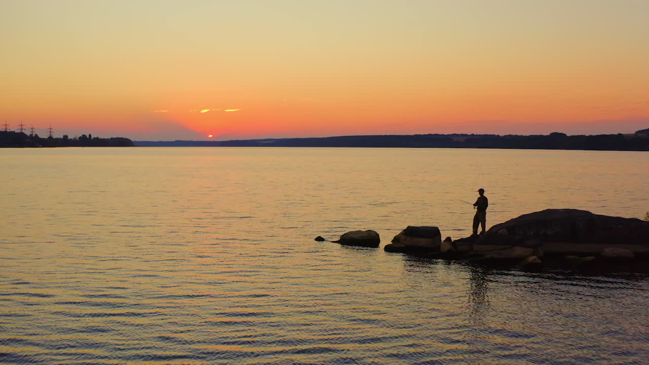 Fisherman at the evening river. Silhouette of a man fishing on the stone in water on the beautiful nature background at sunset.
