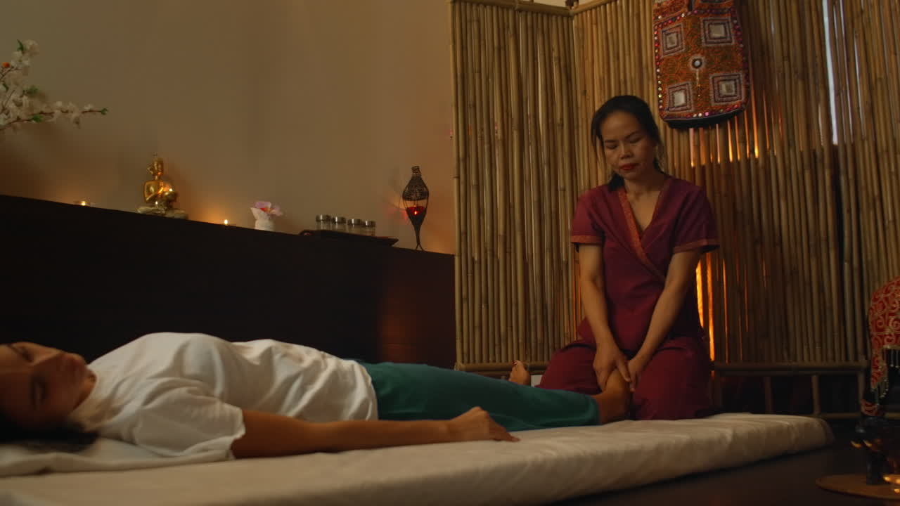 Asian performs massage in national clothes of Thailand. Asian Spa salon in Europe. Young Caucasian woman relaxes while performing massage.