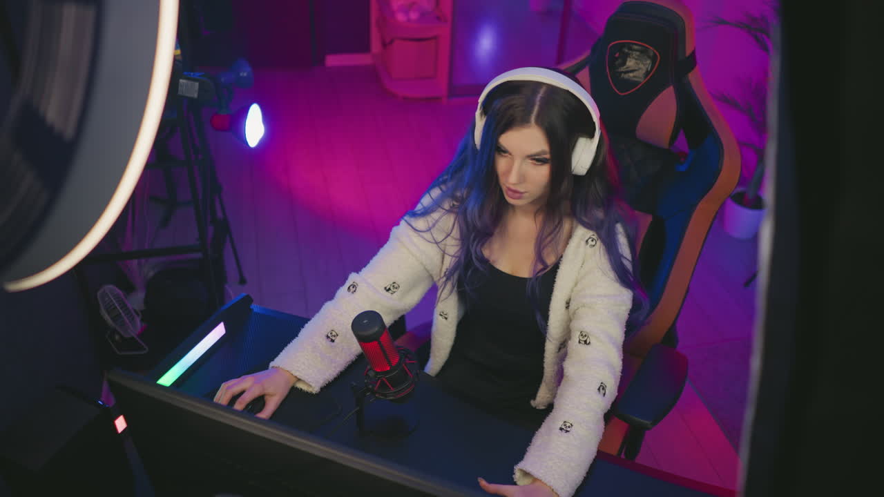 aerial view of pensive girl wearing white headphones sitting in gaming chair looking surprised while playing video game at night in cozy neon studio with condenser mic and dual monitor setup