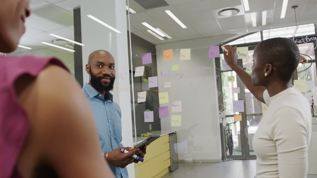 Happy african american colleagues brainstorming, making notes on glass wall in office in slow motion