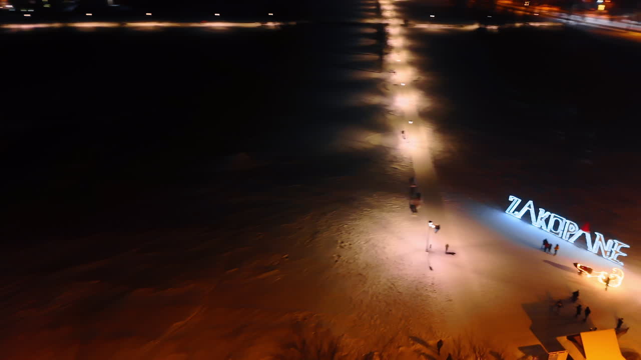 Snow-covered square with Zakopane sign with bright illumination. People wave to the camera. Aerial perspective on the town at night.