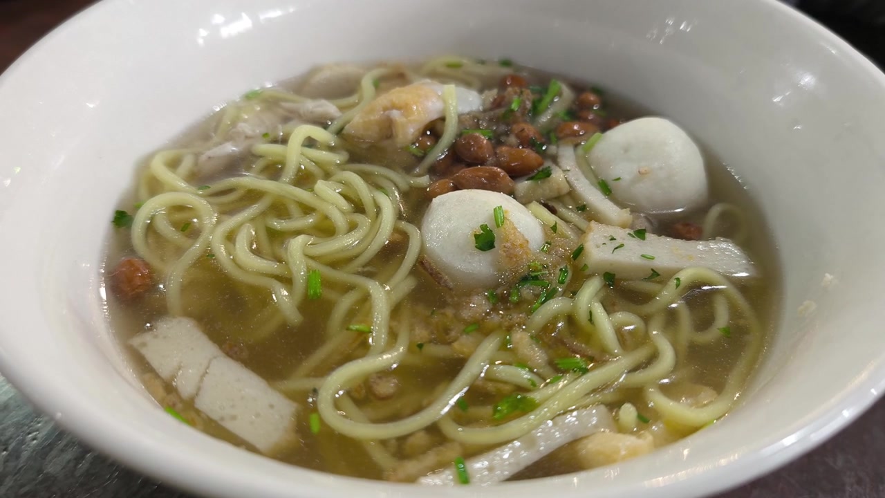 Zuppa di noodle asiatica servita calda in una ciotola di porcellana bianca. Noodle gialli con polpette di pesce, tortini di pesce e verdure miste in un brodo leggero e trasparente. Presentazione del pasto fresca e invitante.