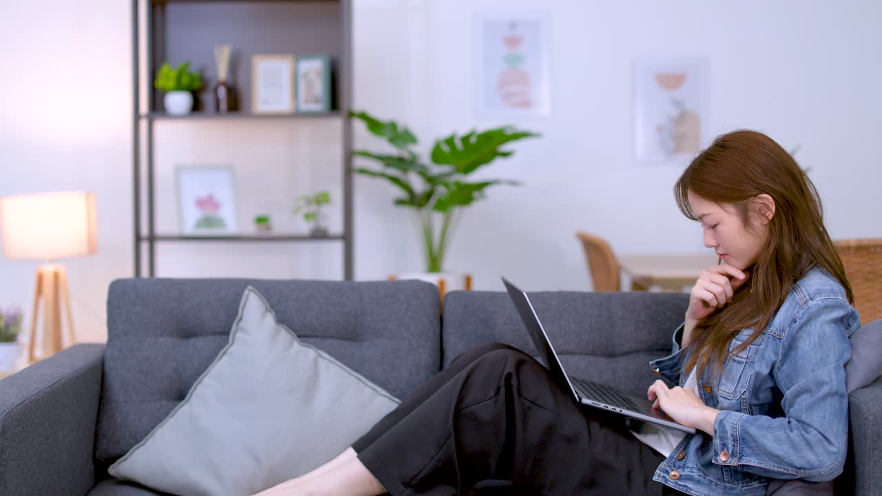 A young Asian woman in a denim jacket types on a laptop while sitting comfortably