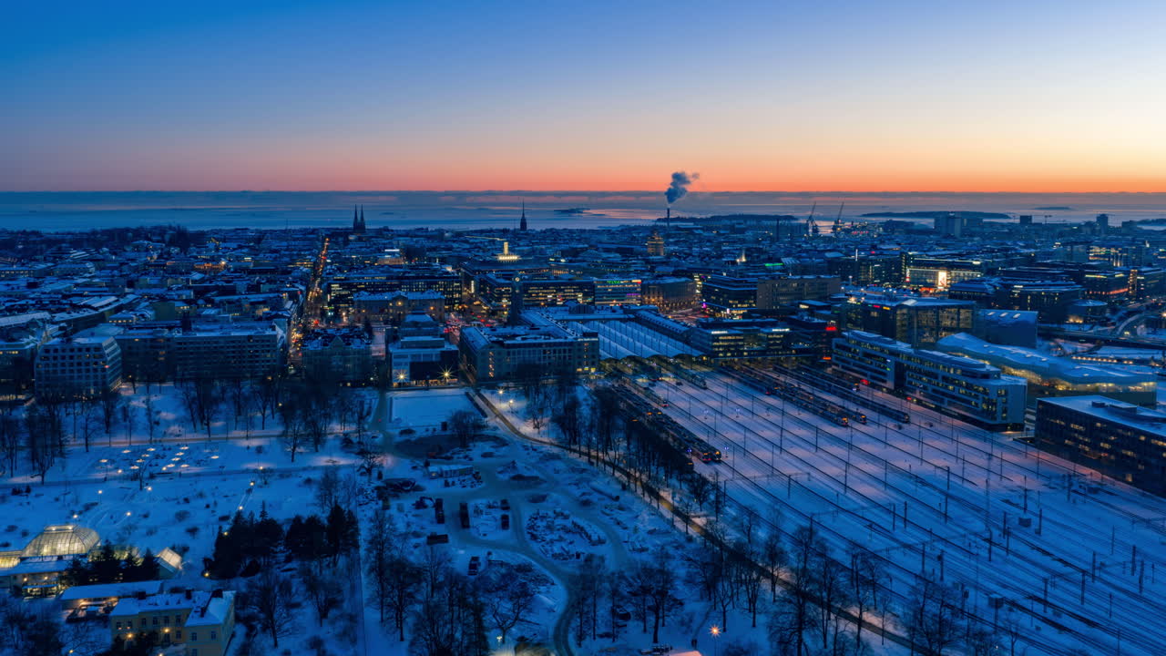 Hyperlapse con drone del Parco Kaisaniemi all'ora blu di Helsinki: movimento laterale sopra il giardino botanico, i treni della stazione ferroviaria, il paesaggio urbano fino al mare ghiacciato. Bagliore crepuscolare invernale, vista innevata. 4K UHD