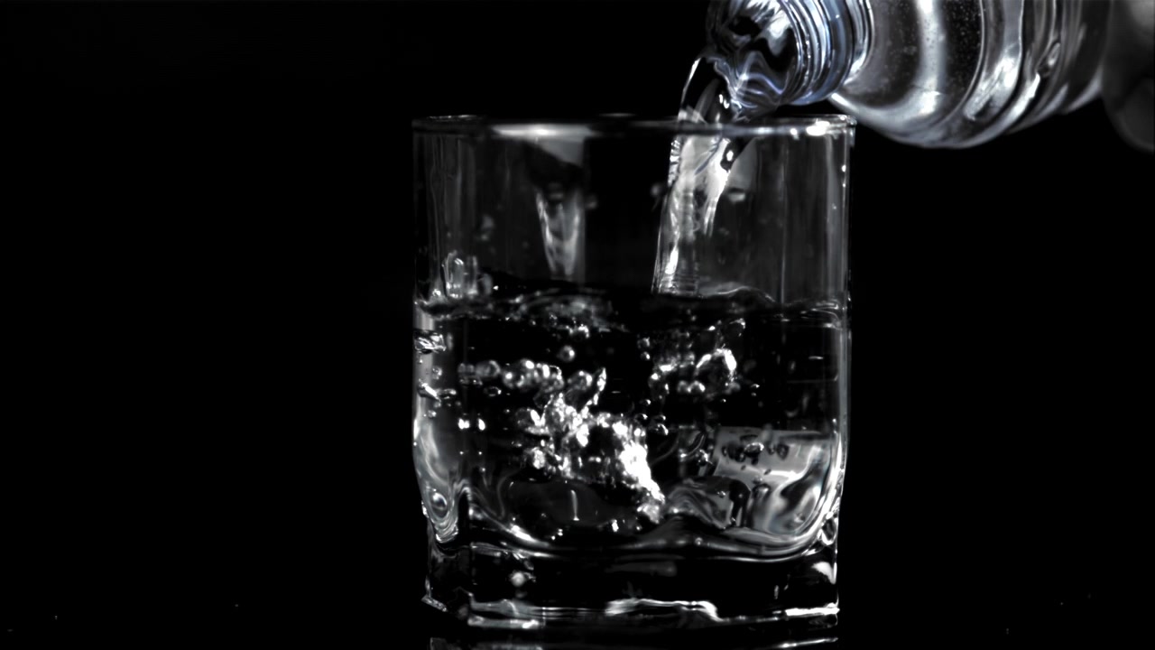 Slow motion-hældning af vand fra flaske i glas på sort baggrund.