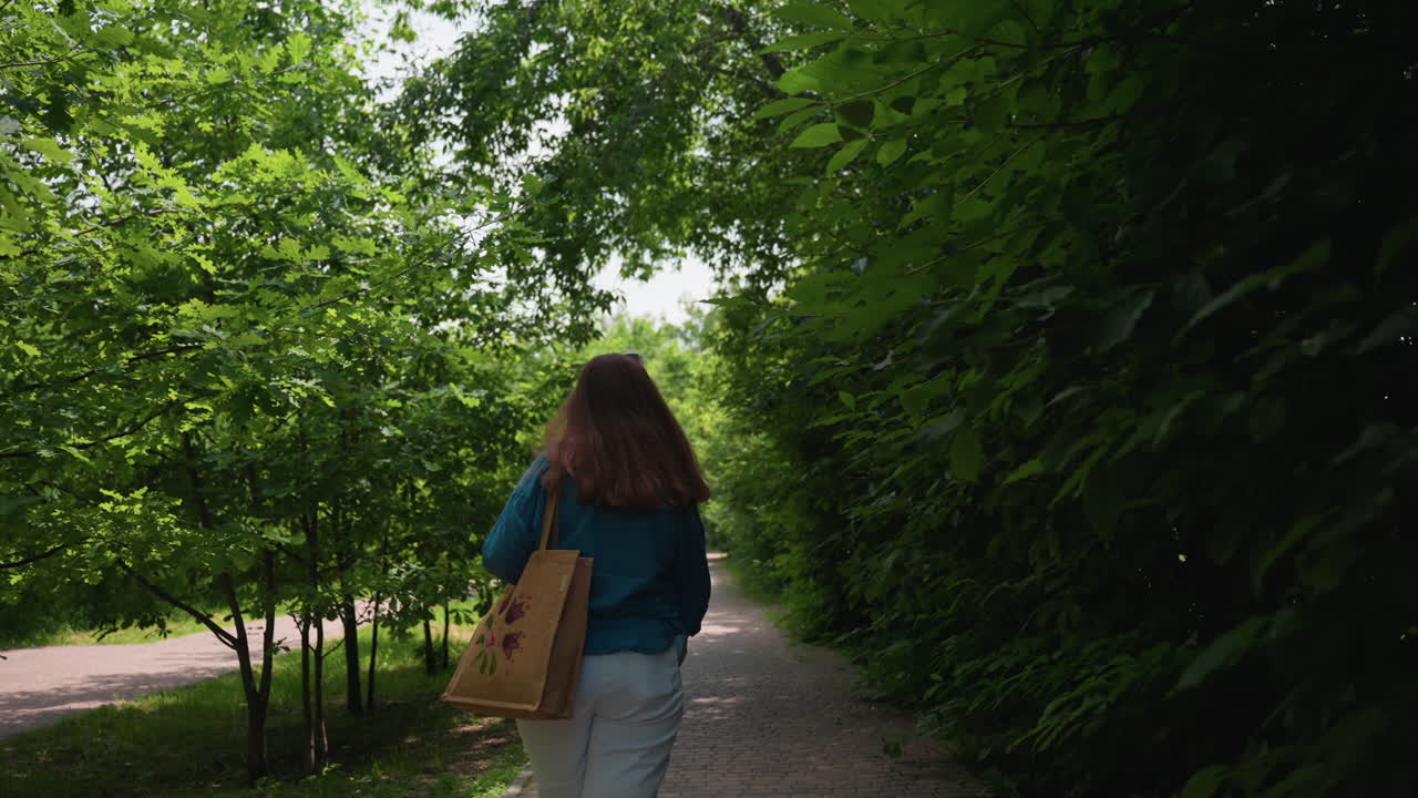 Rear view of lady wearing blue shirt and white shorts carrying handbag walking along quiet park alley surrounded by lush green trees, enjoying peaceful summer day with soft sunlight