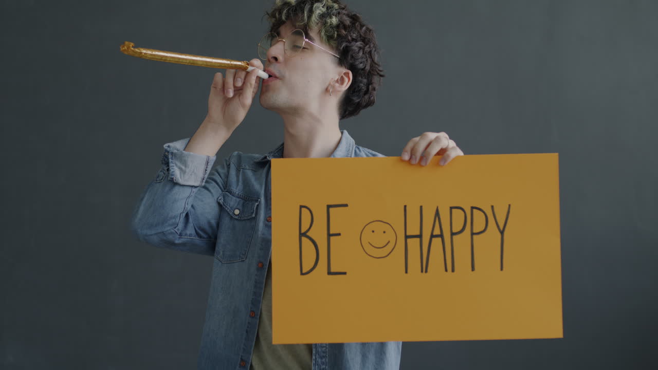 Man holding a sign that says 'Be Happy' while blowing a party horn