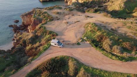 aerial view circling campervan parked on praia dos arrifes rocky coastal cliff on portuguese south coast tilting to reveal resort travel destination