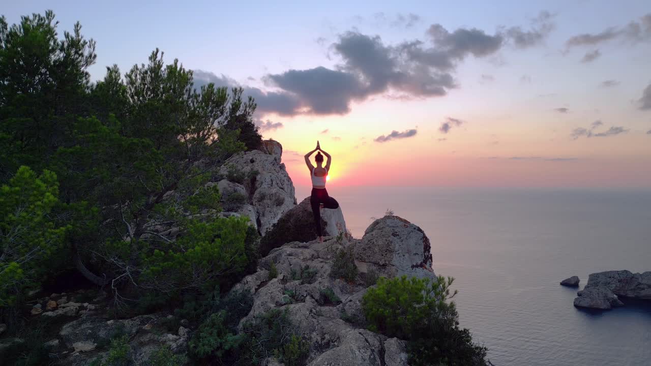 Spettacolare ripresa aerea dall'alto: scogliera di Ibiza, modella in posizione yoga dell'albero al tramonto serale.