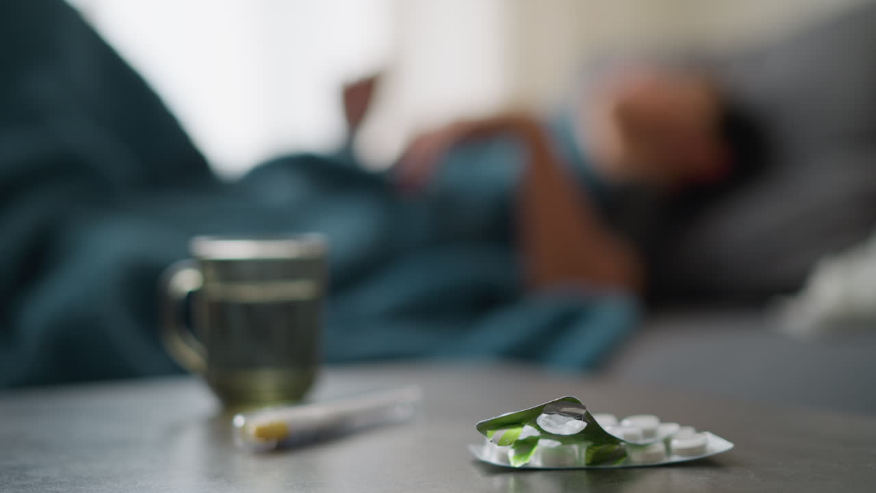Close-up of medication and glass cup on table with blurred view of individual adjusting on bed, suggestive of sick day, recovery, and comfort in a peaceful indoor environment