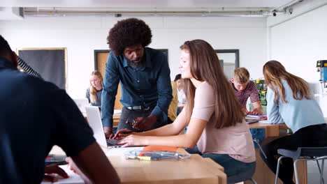 teacher talking to female high school student sitting at work bench using laptop in design and technology lesson