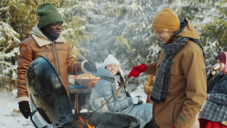 men eating and chatting by bbq grill on winter picnic in woods